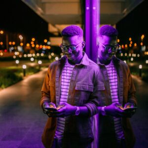 A man in purple lighting leaning against a window and checking his phone