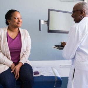 A doctor speaks to a patient in an examination room. Both are smiling.